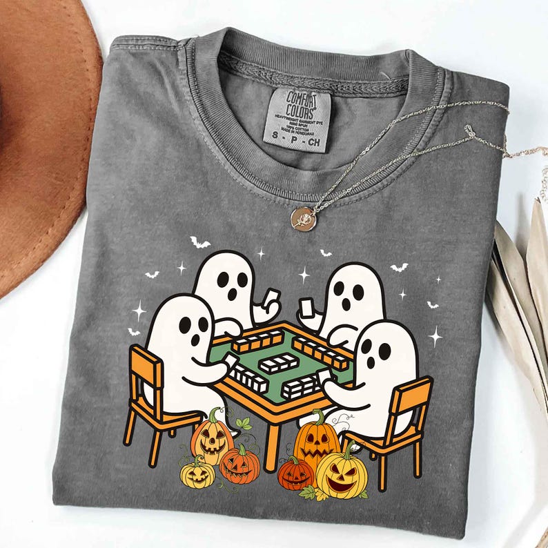 May include: Gray Comfort Colors t-shirt featuring a Halloween-themed graphic. The design shows four cartoon ghosts playing a game at a table, surrounded by pumpkins. A silver necklace with a pendant is draped across the shirt.