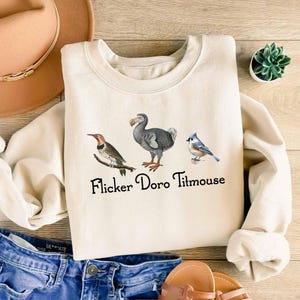 Pode incluir: Uma camisa de moletom cor creme com um gráfico de três pássaros: um dodô, um pássaro azul e um pica-pau. O texto "Flicker Doro Titmouse" é impresso abaixo dos pássaros.