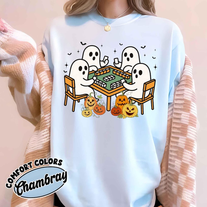 May include: Light blue sweatshirt with a Halloween-themed graphic. The design features four white ghosts playing a game at a table, surrounded by jack-o'-lanterns. The text "COMFORT COLORS Chambray" is printed on the bottom left.