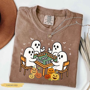 May include: Espresso-colored Comfort Colors t-shirt featuring a Halloween-themed graphic. The design shows four white ghosts playing a game at a green table, surrounded by jack-o'-lanterns. The shirt has a crew neck and short sleeves. A silver necklace is draped across the shirt.