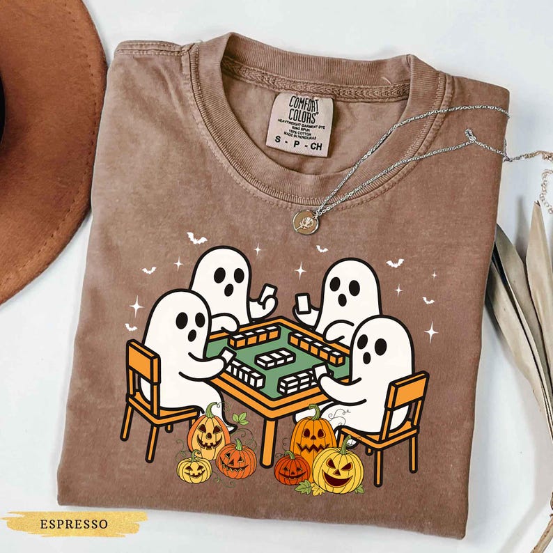May include: Espresso-colored Comfort Colors t-shirt featuring a Halloween-themed graphic. The design shows four white ghosts playing a game at a green table, surrounded by jack-o'-lanterns. The shirt has a crew neck and short sleeves. A silver necklace is draped across the shirt.