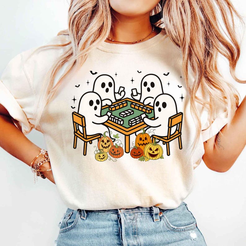 May include: Beige t-shirt featuring a Halloween-themed graphic. The design shows five cartoon ghosts playing a game at a table, surrounded by pumpkins. The ghosts are white with black eyes, and the table is orange with green game pieces.