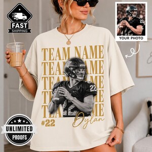 May include: A cream-colored t-shirt featuring a large graphic of a football player in a helmet holding a ball. The text "TEAM NAME" is repeated in gold behind the player, with "#22" and "Dylan" printed below. The shirt has a loose, boxy fit.