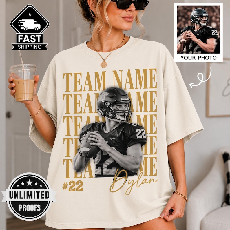 May include: A cream-colored t-shirt featuring a large graphic of a football player in a helmet holding a ball. The text "TEAM NAME" is repeated in gold behind the player, with "#22" and "Dylan" printed below. The shirt has a loose, boxy fit.