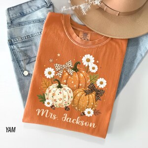May include: An orange t-shirt featuring a graphic of three pumpkins, white daisies, and leopard print bows. The pumpkins are decorated with floral patterns and the text Mrs. Jackson is printed in white script below the design. The shirt is layered with blue denim jeans and a tan hat.