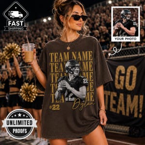 May include: A dark brown oversized t-shirt featuring a graphic print of a football player in a helmet holding a ball, with the text TEAM NAME repeated in gold, the number 22, and a script font. The shirt has a vintage wash finish.