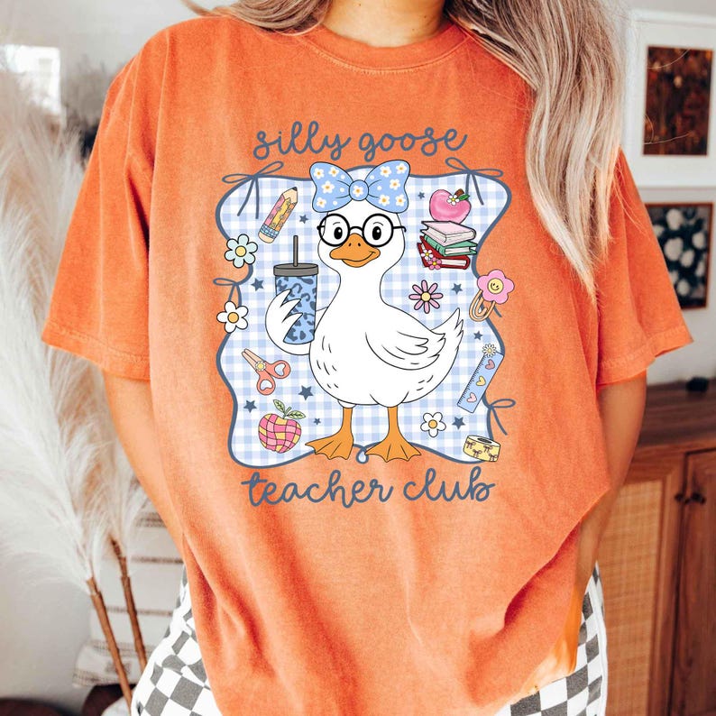K&ouml;nnte beinhalten: Orangefarbenes T-Shirt mit einer Cartoon-Gans, die eine Brille tr&auml;gt und ein Getr&auml;nk h&auml;lt. Das Design enth&auml;lt den Text "silly goose teacher club" mit Illustrationen zum Thema Schule.