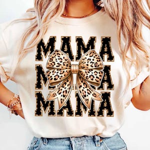 Puede incluir: Camiseta blanca con la palabra "MAMA" impresa tres veces en negro con un contorno dorado. Un gran lazo de estampado de leopardo está centrado entre las palabras.