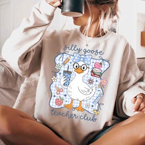 K&ouml;nnte beinhalten: Ein hellbeige Sweatshirt mit einer Grafik einer wei&szlig;en Gans mit Brille und einer blauen Schleife. Die Gans h&auml;lt ein Getr&auml;nk und ist von Illustrationen zum Thema Schule umgeben. Der Text "silly goose teacher club" steht auf dem Sweatshirt.