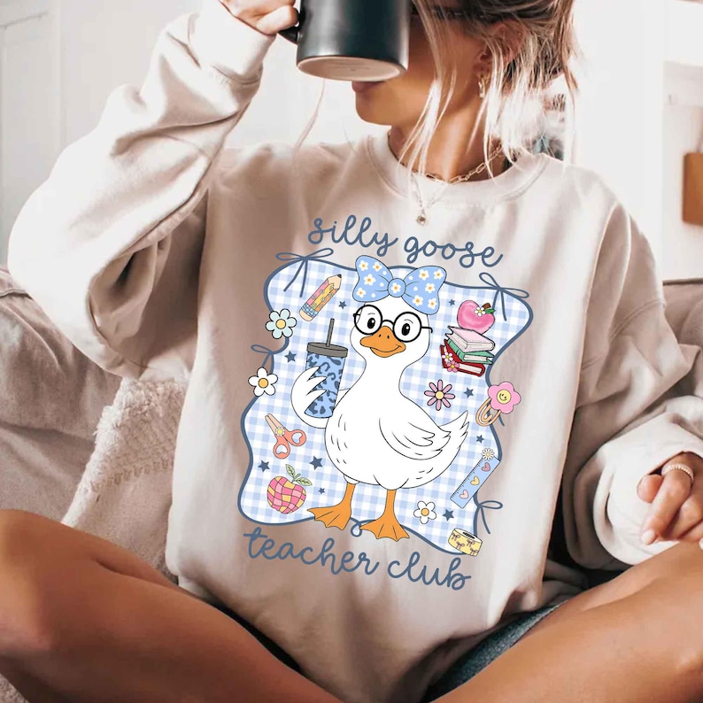 K&ouml;nnte beinhalten: Ein hellbeige Sweatshirt mit einer Grafik einer wei&szlig;en Gans mit Brille und einer blauen Schleife. Die Gans h&auml;lt ein Getr&auml;nk und ist von Illustrationen zum Thema Schule umgeben. Der Text "silly goose teacher club" steht auf dem Sweatshirt.
