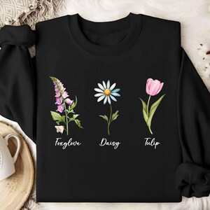 Puede incluir: Sudadera negra con un gráfico de tres flores: un tulipán rosa, una margarita blanca y una dedalera morada. Las flores están etiquetadas con sus nombres.