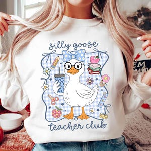 K&ouml;nnte beinhalten: Wei&szlig;es Sweatshirt mit einer Cartoon-Gans, die eine Brille und eine Schleife tr&auml;gt. Die Gans h&auml;lt ein Getr&auml;nk und ist von schulbezogenen Grafiken umgeben, darunter Bleistifte, B&uuml;cher und Blumen. Der Text "silly goose teacher club" ist auf dem Sweatshirt aufgedruckt.