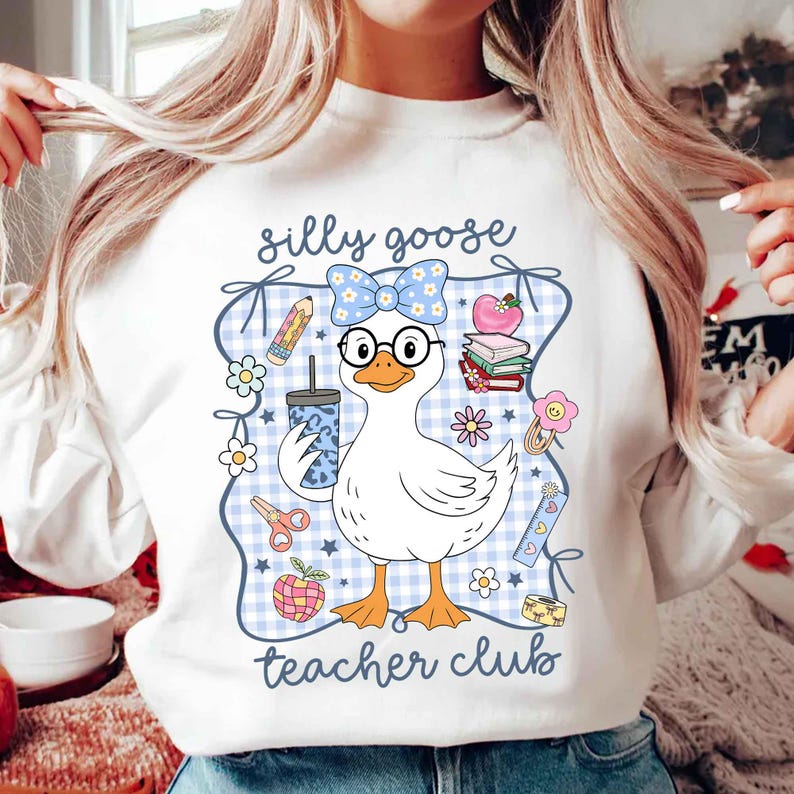 K&ouml;nnte beinhalten: Wei&szlig;es Sweatshirt mit einer Cartoon-Gans, die eine Brille und eine Schleife tr&auml;gt. Die Gans h&auml;lt ein Getr&auml;nk und ist von schulbezogenen Grafiken umgeben, darunter Bleistifte, B&uuml;cher und Blumen. Der Text "silly goose teacher club" ist auf dem Sweatshirt aufgedruckt.