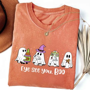 May include: Orange t-shirt with a crew neck featuring a Halloween-themed graphic. The graphic shows four cartoon ghosts, each with eye-related accessories, and the text "Eye see you, Boo". A gold necklace is draped on the shirt.