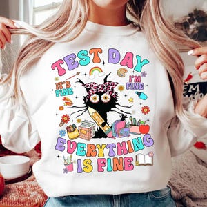 May include: White sweatshirt with a colorful graphic that reads "TEST DAY EVERYTHING IS FINE!" The design features a black cat with a leopard print headband, a pencil, books, and school supplies.