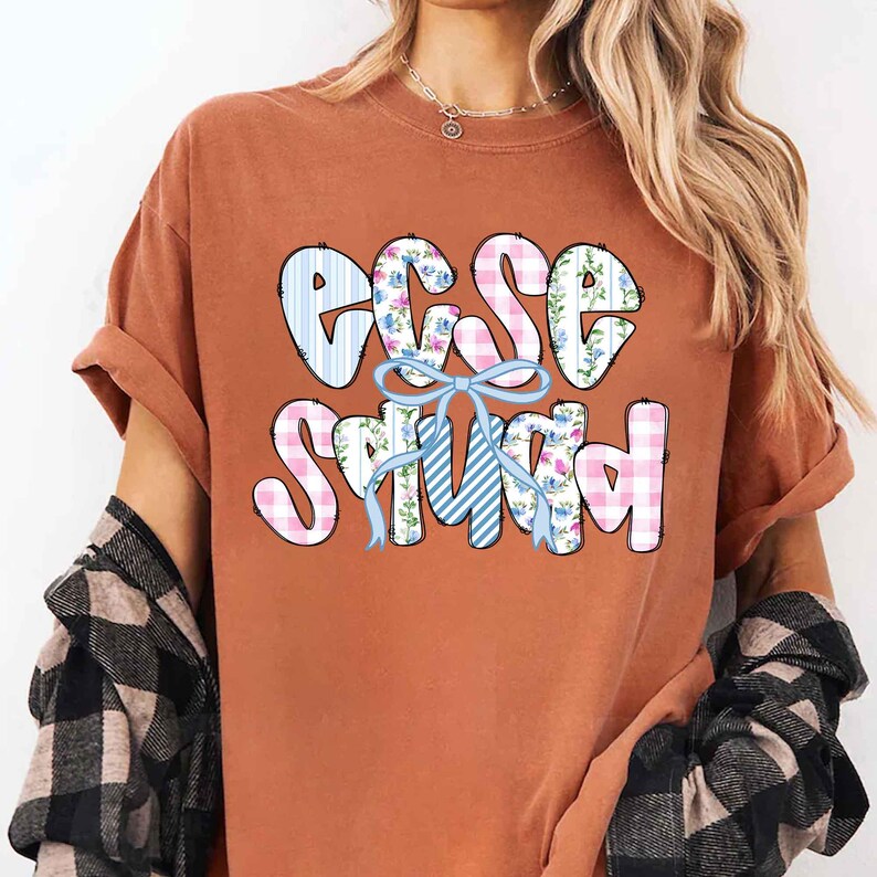 Puede incluir: Camiseta naranja quemado con la frase "ECSE Squad" en una fuente l&uacute;dica. Las letras est&aacute;n decoradas con rayas azules y blancas, cuadros de vichy rosas y estampados florales, con un lazo azul claro en el centro.