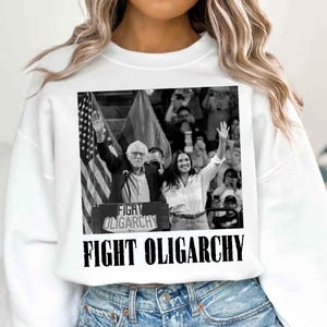 Puede incluir: Una sudadera blanca con una foto en blanco y negro de dos personas levantando las manos al aire. La foto es de Bernie Sanders y Alexandria Ocasio-Cortez. El texto "FIGHT OLIGARCHY" está impreso en negro en la sudadera.