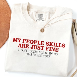 May include: Cream-colored t-shirt with the text "MY PEOPLE SKILLS ARE JUST FINE, IT'S MY TOLERANCE TO IDIOTS THAT NEEDS WORK" in red. A gold necklace with a pendant is draped across the shirt. The shirt is folded.