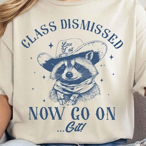 Może przedstawiać: Biały t-shirt z niebieskim nadrukiem przedstawiającym szop pracza w kowbojskim kapeluszu i bandanie. Tekst "Class Dismissed" znajduje się nad szop praczem, a "Now Go On...Git!" pod szop praczem. Kapelusz szop pracza ma napis "Love Y'all".