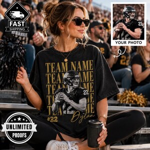 May include: A black oversized t-shirt featuring a graphic of a football player in a helmet holding a ball, with the repeated text "TEAM NAME" in gold, the name "Dylan" in script, and the number "22" printed on the front of the garment.