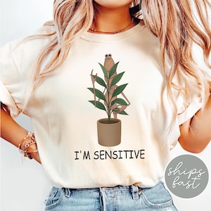 May include: Cream-colored sweatshirt with a graphic of a plant-like figure in a pot, holding a stick. The text "I'M SENSITIVE" is printed below the graphic. The sweatshirt is a casual, comfortable style.