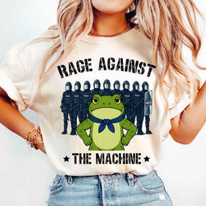 May include: Beige t-shirt with the text "RAGE AGAINST THE MACHINE" above a cartoon frog wearing a blue scarf, standing in front of a line of police officers in riot gear. The shirt has a crew neck and short sleeves.