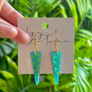 Puede incluir: Un par de pendientes colgantes geométricos. Los pendientes tienen forma triangular, en color azul turquesa con detalles de triángulos verdes y ganchos dorados. Se exhiben en una tarjeta beige con el texto "Lazy Autumn".