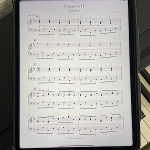 May include: A digital tablet displaying sheet music for Pachelbel's Canon in D, resting on a piano. The music is in black and white, with the title and composer's name visible at the top. The tablet is in a black case.