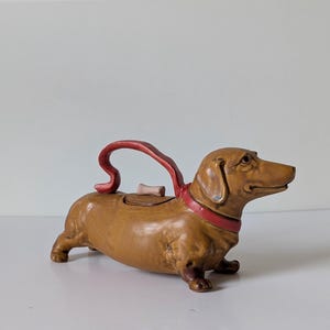 Puede incluir: Una tetera de cerámica marrón con forma de perro salchicha. La tetera presenta un collar rojo que se extiende hasta formar un asa roja curvada en la parte posterior. Un pequeño pomo de color claro con forma de hueso se sitúa en la tapa.