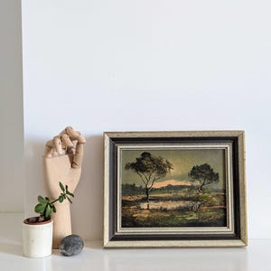 Op de afbeelding: Vintage landschapsschilderij in een houten lijst. Het kunstwerk toont twee bomen in een veld met water, onder een zonsondergang. De lijst is donkerbruin en crème. Een kleine potplant en een houten hand staan naast het schilderij.