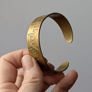 Op de afbeelding: Een goudkleurige manchetarmband met een gegraveerd geometrisch patroon. De open armband wordt in een hand gehouden tegen een neutrale achtergrond. De armband heeft een licht gebogen vorm.
