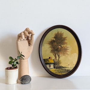 Op de afbeelding: Een ovaalvormig schilderij met een landschap met een huis, boom en hek. De lijst is donkerbruin. Een houten gearticuleerde hand, een kleine potplant en een grijze steen zijn ook in de afbeelding te zien.