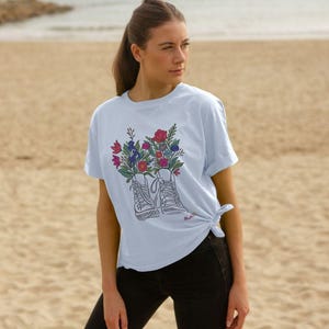 May include: Pale blue t-shirt with a graphic of trainers sprouting colorful flowers. The illustration shows red, pink, blue, and purple blooms. The shirt is knotted at the waist and is worn with black trousers.