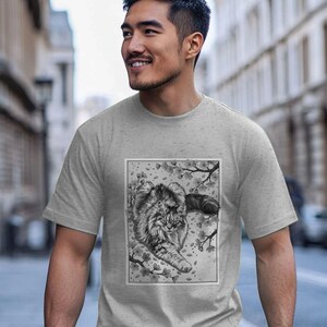 Könnte beinhalten: Ein hellgraues, meliertes Kurzarm-T-Shirt mit einem rechteckigen Grafikdruck auf der Brust. Der Druck zeigt eine langhaarige Katze, die auf einem Ast liegt, umgeben von Kirschblüten in einem detaillierten schwarz-weißen Zeichenstil.
