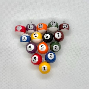 May include: A set of miniature snooker balls, each with a different colour and number from 1 to 15. The balls are arranged in a triangle, against a white backdrop. The balls are small and appear to be ornaments.
