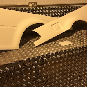 May include: Two white fibreglass car body panels, probably for a custom car project, are shown on a brown diamond-patterned surface.