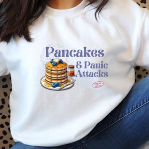 Może przedstawiać: Biała bluza z nadrukiem graficznym stosu naleśników z borówkami i syropem oraz tekstem "Pancakes & Panic Attacks" w fioletowym kolorze. Tekst "Keep calm and get some" jest w kolorze czerwonym.