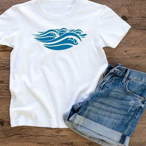 May include: White t-shirt with a blue wave design and denim shorts. The t-shirt features a stylized wave graphic in the center. The shorts are blue denim with rolled-up cuffs, resting on a wooden surface.