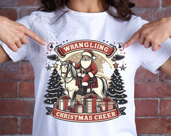 May include: White t-shirt with a Christmas-themed design featuring Santa Claus riding a horse with the text "Wrangling Christmas Cheer".