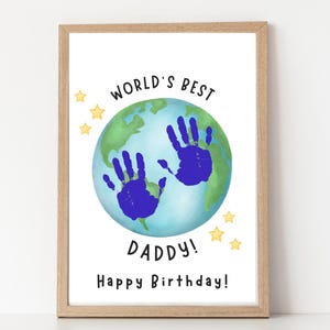 Puede incluir: Una impresión enmarcada con el texto "WORLD'S BEST DADDY! Happy Birthday!". La obra de arte presenta un globo azul y verde con dos huellas de manos azules. Estrellas amarillas están dispersas alrededor del globo.
