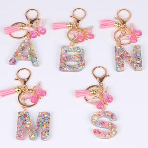 Könnte beinhalten: Fünf Schlüsselanhänger mit den Buchstaben A, B, N, M und S. Jeder Buchstabe ist mit bunten Glitzer gefüllt und hat einen goldfarbenen Schlüsselring, eine rosa Quaste und einen Schmetterlingsanhänger. Die Schlüsselanhänger sind vor weißem Hintergrund.