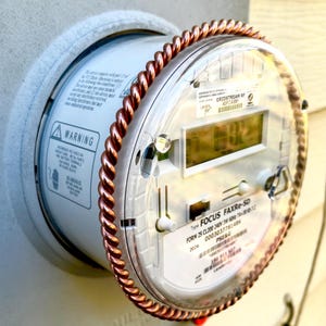 May include: A round digital electricity meter with a clear plastic cover and a copper-colored twisted metal ring around the edge. The device features a digital display, a warning label, and text including Gridstream RF, Focus FAXRe-SD, and PSE&G.