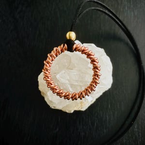 May include: A copper-colored wire-wrapped pendant necklace. The pendant is a circular design with a black cord and a gold-colored bead. The pendant rests on a white crystal against a black background.