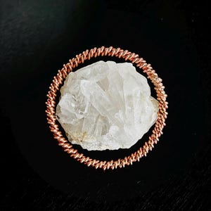May include: A copper-colored wire bracelet encircles a large, clear quartz crystal. The bracelet is made of tightly wound wire, creating a textured, circular design. The crystal is rough and irregular in shape, with multiple facets.