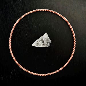 May include: A copper-colored, twisted rope necklace encircles a clear, irregularly shaped crystal on a dark surface. The necklace is circular, and the crystal is positioned in the center. The overall aesthetic is minimalist and natural.