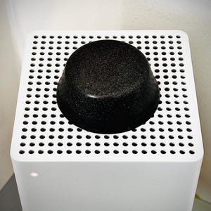 May include: A black, rounded object with a textured surface sits atop a white, perforated square. The object is centered on the square, which is mounted on a white, rectangular base. The overall aesthetic is minimalist and modern.