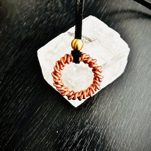 May include: A circular pendant crafted from twisted copper wire, suspended from a black cord. A single smooth, matte gold-toned bead sits directly above the copper ring, securing it to the cord. The design features a continuous, uniform coil.