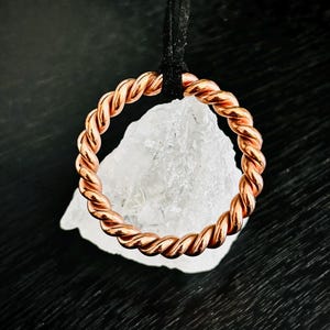 May include: A circular pendant made of twisted copper wire suspended from a black cord. The copper ring encircles a jagged, translucent white quartz crystal. The metallic surface of the copper reflects light, showing a warm, reddish-brown tone.