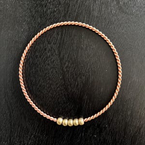 May include: A copper-colored, twisted metal bangle bracelet with five small, gold-colored beads. The bracelet is circular and sits on a dark, textured surface. The twisted design gives the bracelet a rope-like appearance.