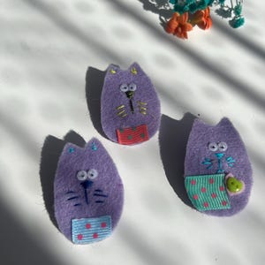 May include: Three handmade purple felt cat-shaped brooches. Each brooch has embroidered details for the eyes, nose, and whiskers. The cats have colourful patterned pockets. Decorative flowers are in the background.